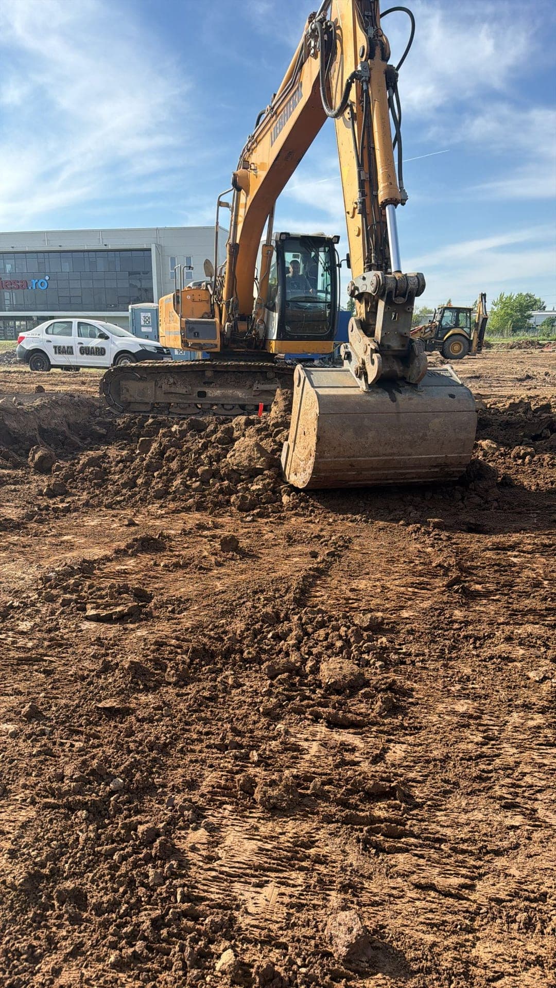 Excavator pe șenile cu cupă în acțiune — excavare strat vegetal pe platforma viitorului magazin Lidl